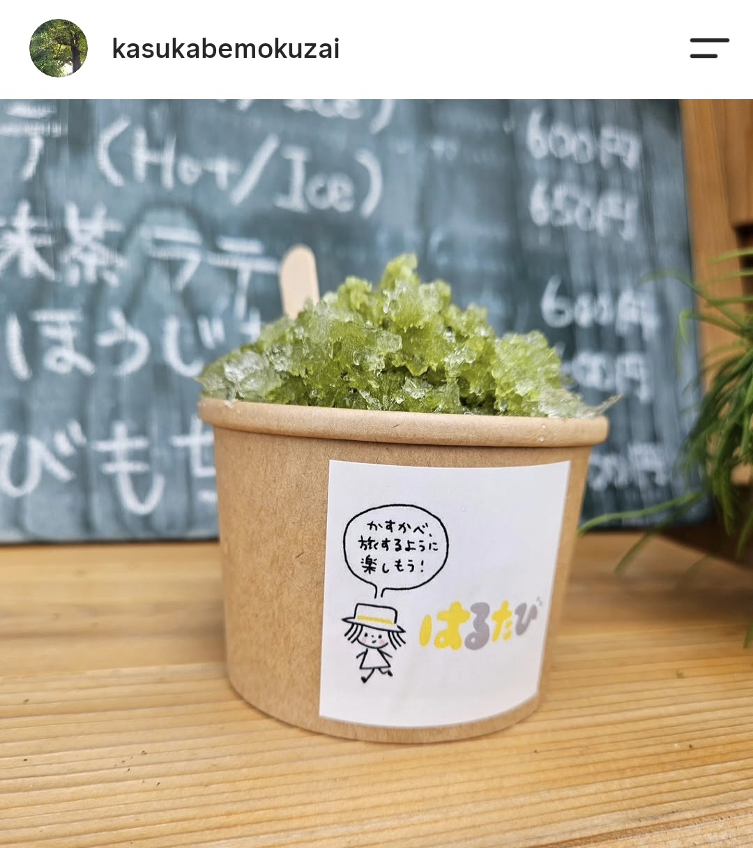 「はるたび」と書かれた茶色の紙コップに抹茶のかき氷を撮影した写真。背景にはメニューが書かれた黒板が写っている。
