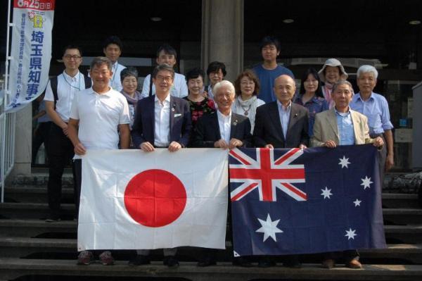 春日部市役所正面入口にある階段にて撮影された春日部市公式訪問団および準公式訪問団と石川市長との記念写真。手前の段に立つ市長や関係者たちは日本とオーストラリアの大きな国旗を並べて持って並んでいる。