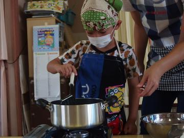 カレー作り中にコンロにのっている鍋の取っ手に手を添えている子供とその横に大人がいる様子の写真