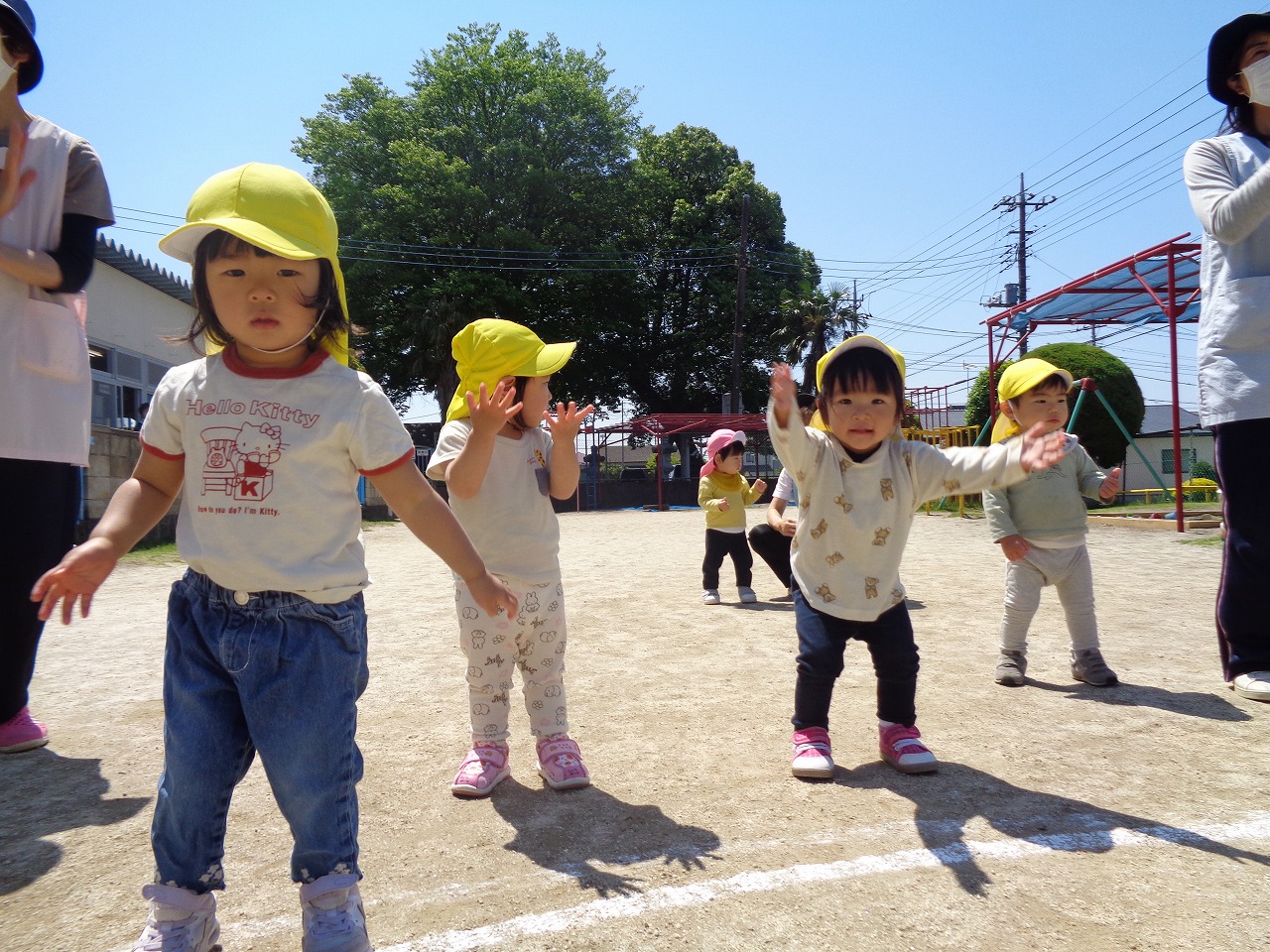 黄色帽子を被った園児達が、保育所の庭で両手を広げ、体を動かして体操している姿の写真