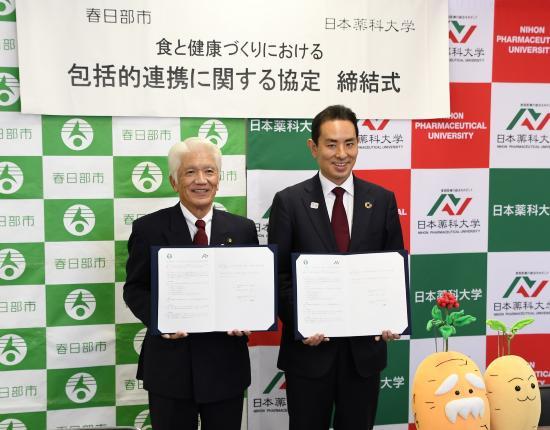 キャラクターのぬいぐるみの横で、二人の男性が笑顔で協定書を持って並んでいる写真