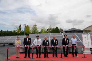 春日部市武里太陽光発電所の竣工式で横一列に並んだ7名の出席者によるテープカット直前の様子を撮影した写真