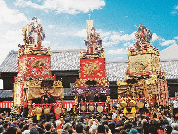 朱塗りの土台に金の飾りや刺繍が施された、豪華絢爛な江戸型人形山車が青空の下担ぎ上げられている写真