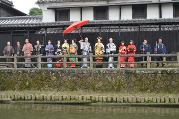 朱塗りの傘を持った人を中央に川沿いに並ぶ着物姿の人々の歌麿まつりの写真