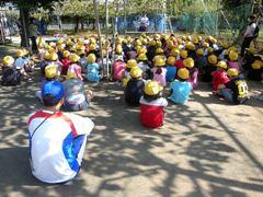 帽子をかぶった児童たちが果樹園で座って説明を受けている写真