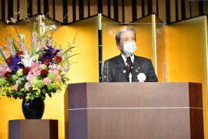 金屏風前の壇上で卓上マイクでアクリル板越しにあいさつをするマスク姿の石川市長と左横の黒色の花瓶に入った色鮮やかな花の写真