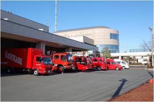春日部市消防本部の建物があり、建物の手前に車庫があり、車庫には救急車1台と消防車5台が並んでいる様子の写真