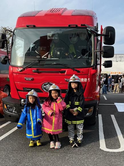 3名のこどもが防火服を着用し消防自動車の前で写真をとる様子