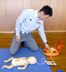 電極パッドをつけた赤ちゃん人形につながっているAEDを操作している男性の写真