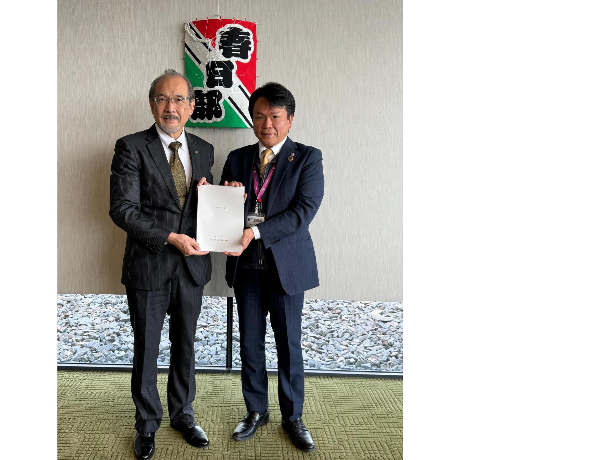 春日部と書かれた凧の前で、審議会会長から答申を受け取る岩谷市長の写真