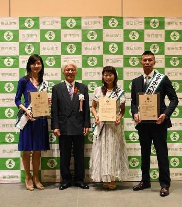 緑色の春日部市のロゴが見える壁面を背に、市長と委嘱状を持つ大使が並んでいる写真