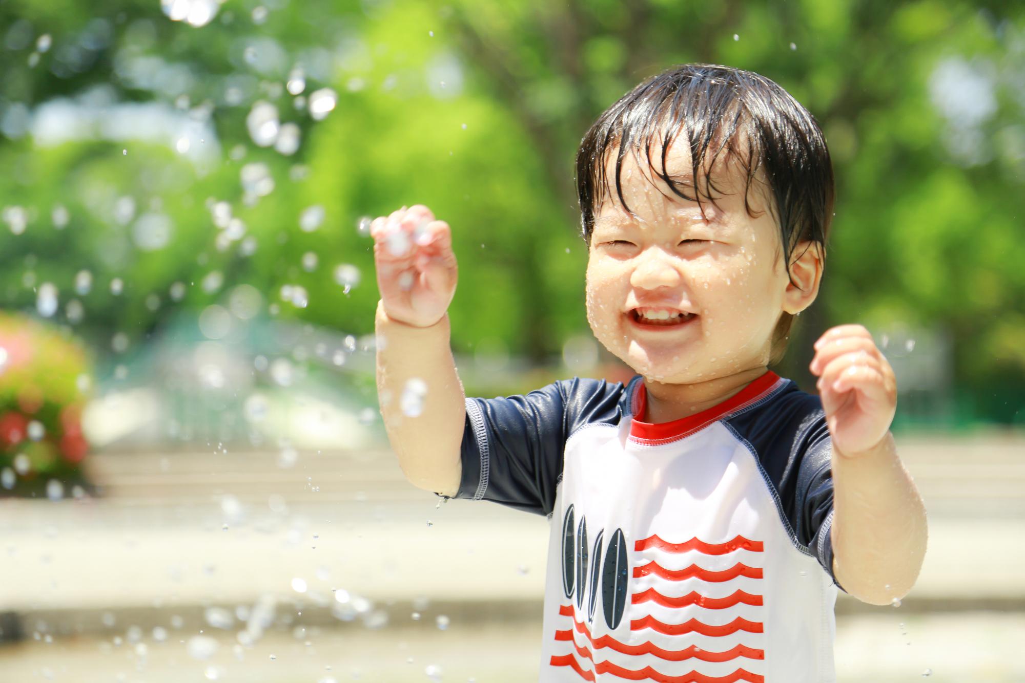 白地に赤色と紺色の模様が入ったTシャツを着た男の子が水遊びをしている様子