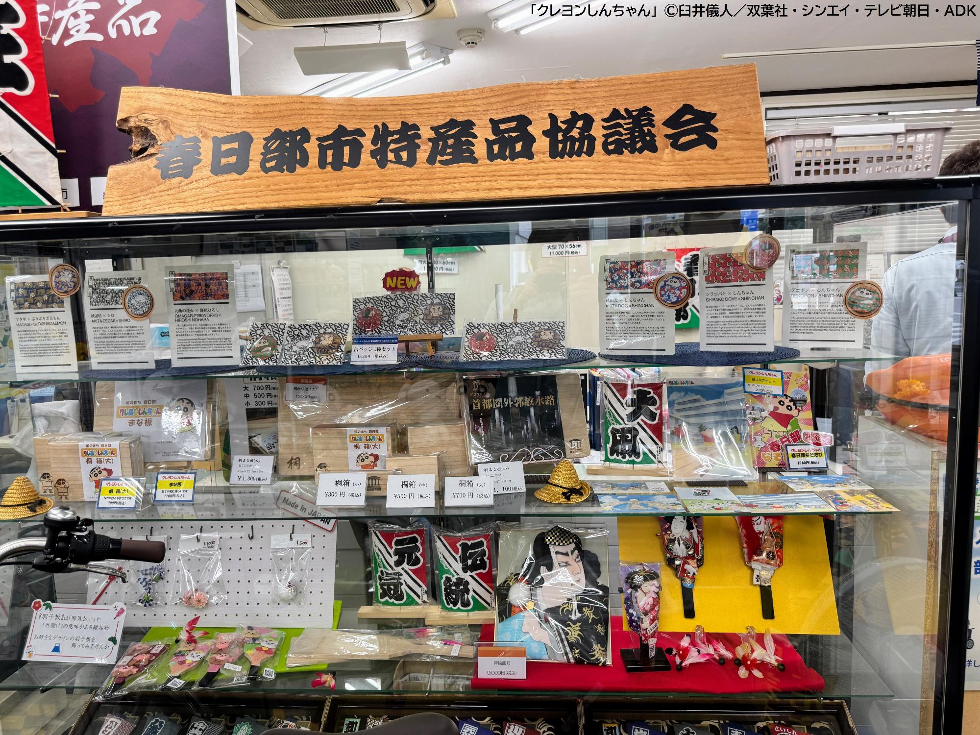 ぷらっとかすかべの施設内にある特産品の紹介を行うガラスケースの写真。押絵羽子板や凧、『クレヨンしんちゃん』の焼き印が入った桐箱や桐のまな板が入れられている。