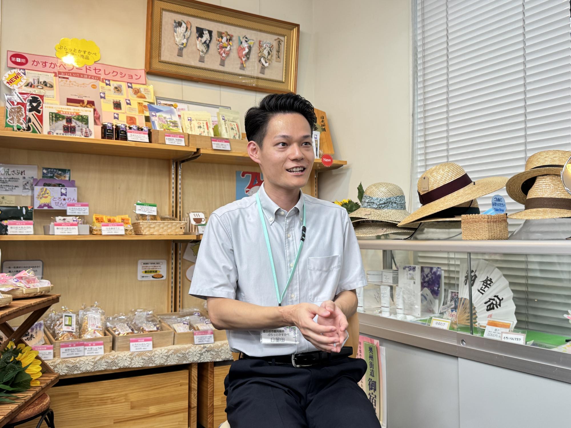 販売しているかすかべフードセレクションや麦わら帽子の前で座り、インタビューに答えている日留田さんの様子。
