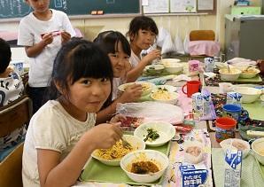 給食を食べている女子児童三名がカメラのほうを向いて笑顔をみせている写真