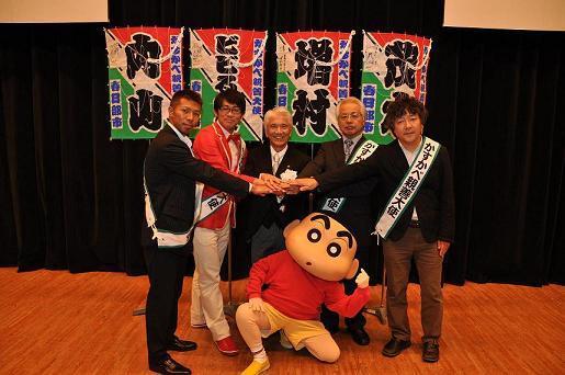 5名の親善大使とクレヨンしんちゃんのキャラクターが手を合わせて記念撮影している写真