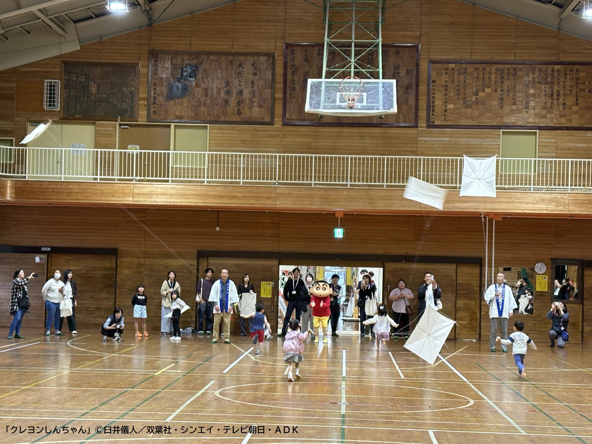 凧あげ体験を楽しむ参加者と、参加者を応援しているしんのすけくん