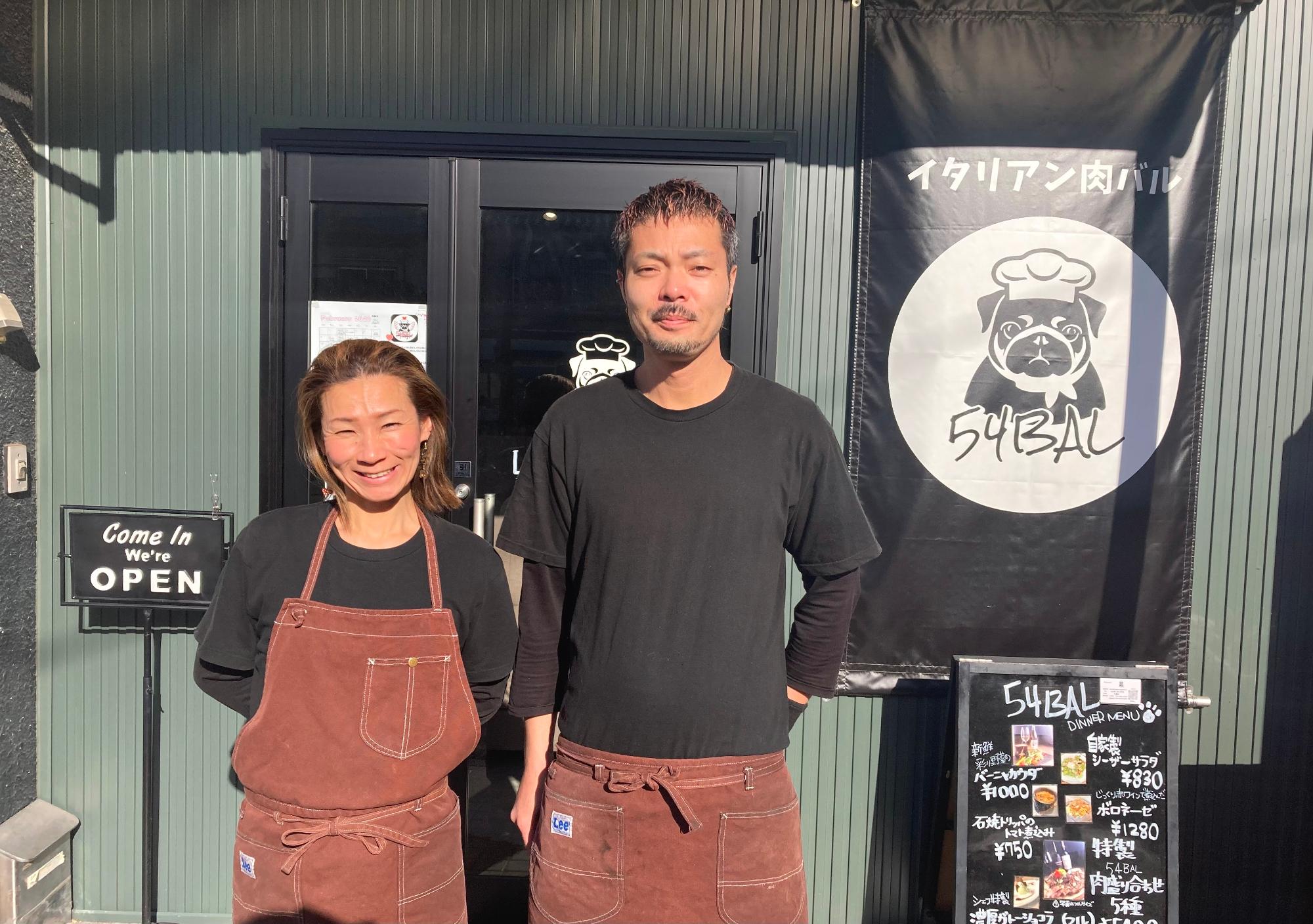 黒いTシャツとえんじ色のエプロンを着て、店舗の前で微笑む五嶌弘太郎さん(右)と真理さん(左)の写真