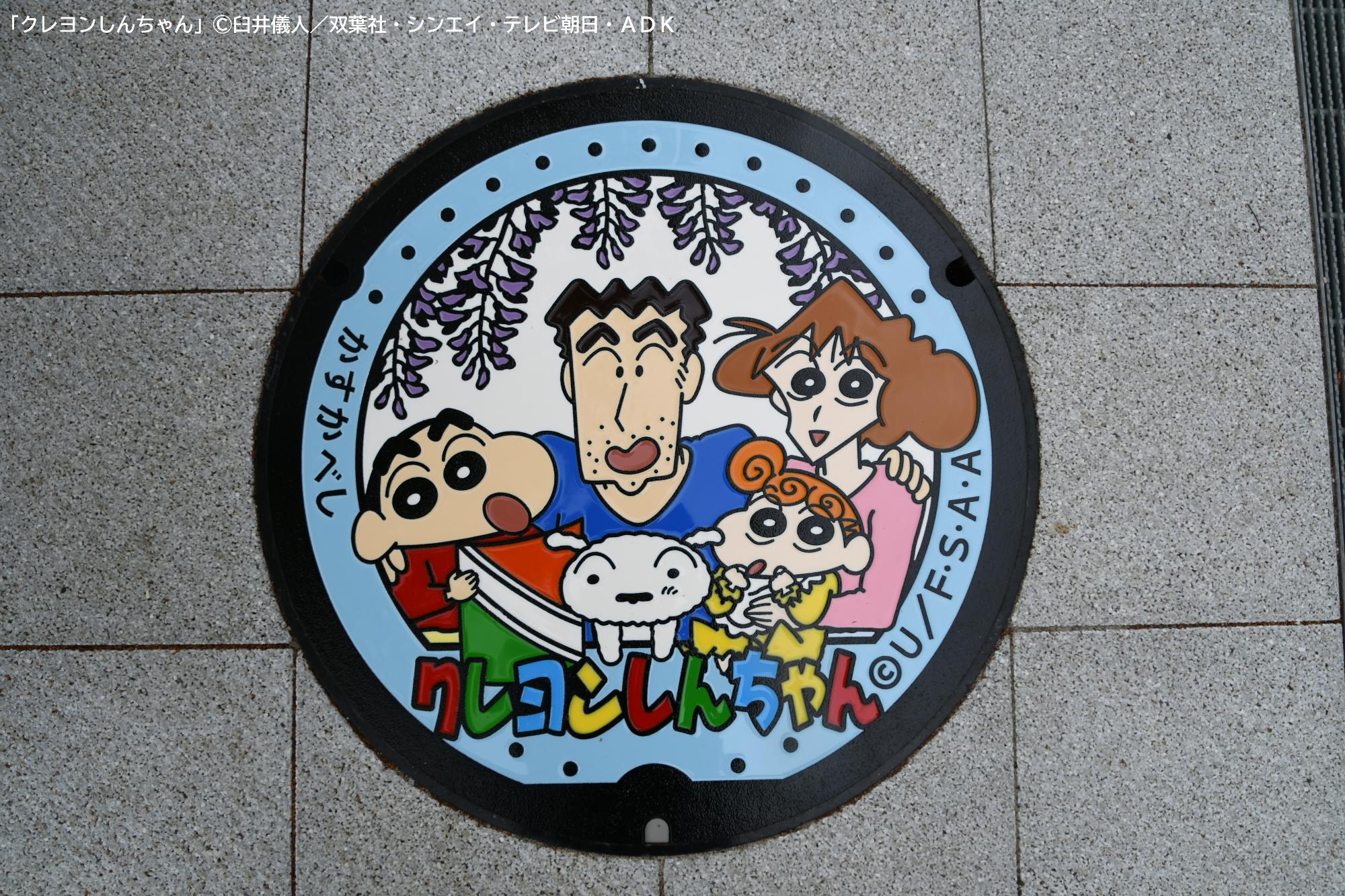 春日部市役所の来庁記念スタンプをイメージしてデザインされたマンホールふたの写真。フジの花の元、『クレヨンしんちゃん』の野原一家の皆さんが笑顔で並んでいる。しんちゃんの手には凧が握られている。