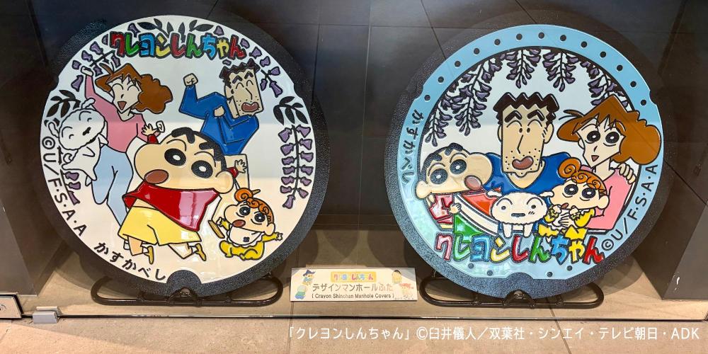 2枚のマンホールふたが展示されている様子。左側が春日部藤まつりで野原一家がパレードしている様子をモチーフにしたデザイン、右側が市役所来庁記念スタンプをモチーフにしたデザイン。