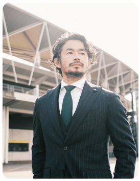 黒いのスーツに緑色のネクタイをして遠くを見ている佐藤 勇人さんの写真