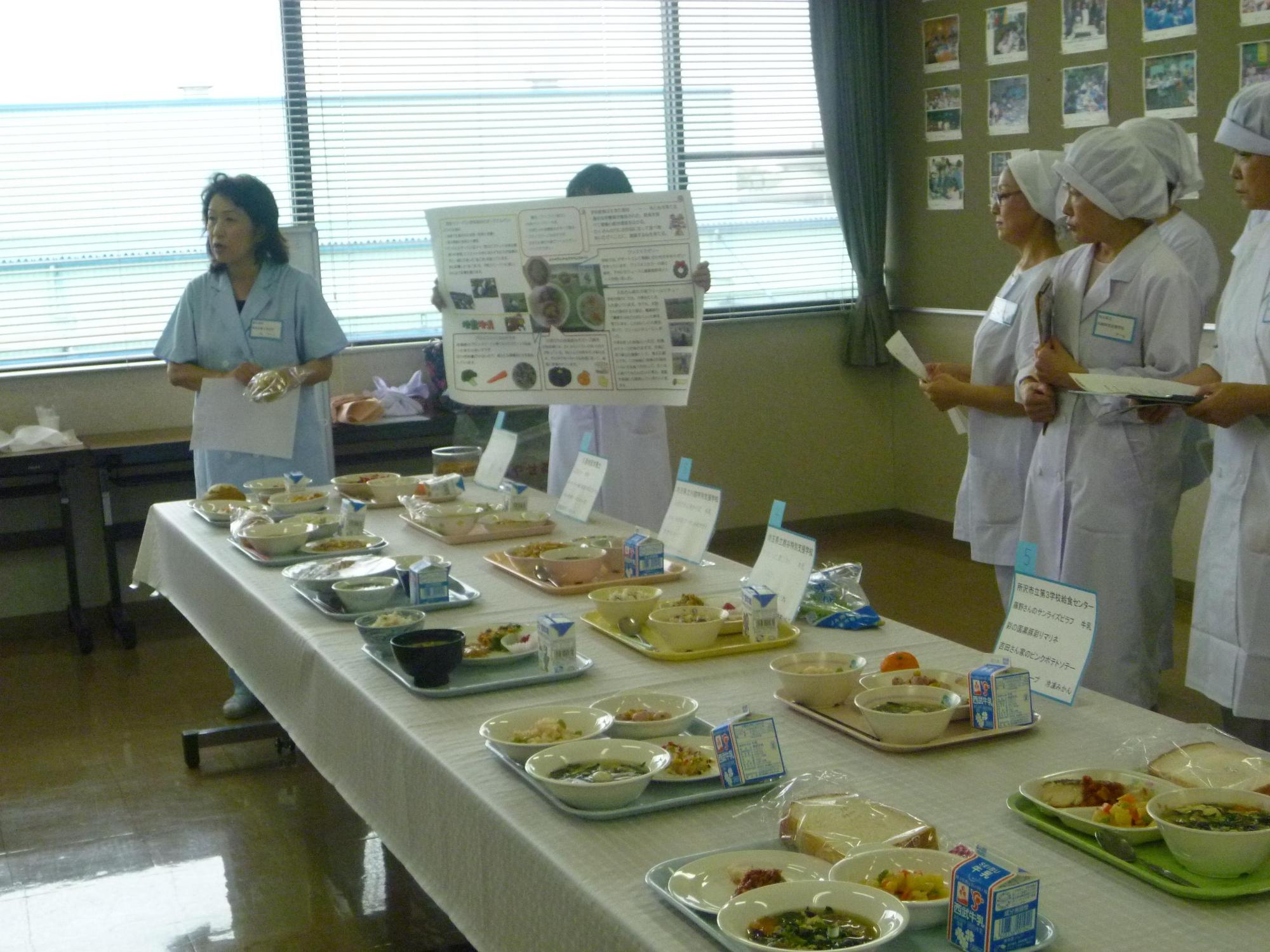 長机の上に10種類以上の献立メニュー別に準備した給食のお盆を並べ話し合う栄養士さんと給食センターの皆さんの写真