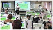 多くの人が並べられたパソコンを見ている前で、男性が立って教鞭を取っている写真